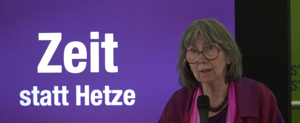Rednerin auf der Bühne vor einem Banner mit der Aufschrift „Zeit statt Hetze“